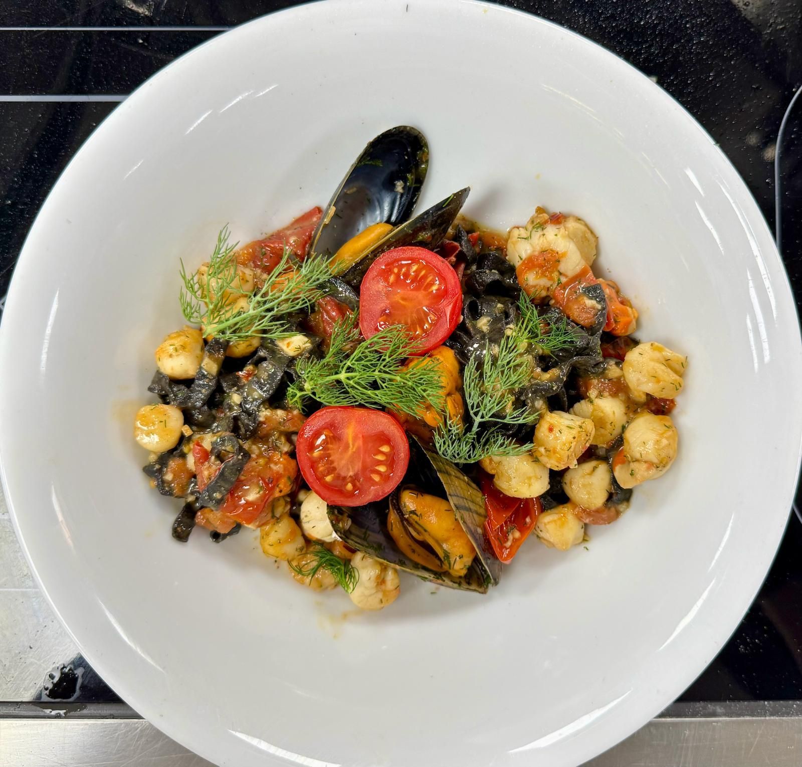 Pasta dish with mussels, scallops, tomatoes, and dill in a white bowl.