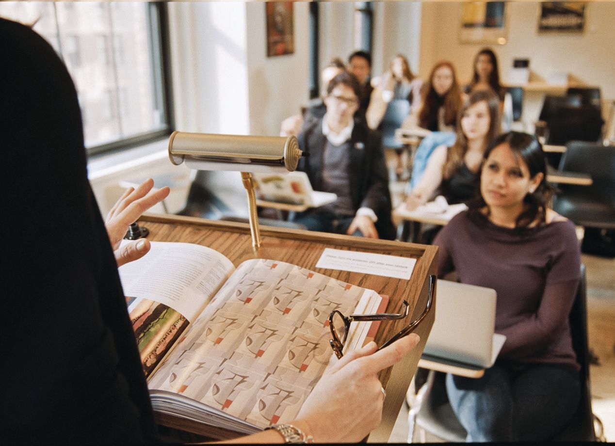 Instructor in black robe lectures from book to students in a classroom.