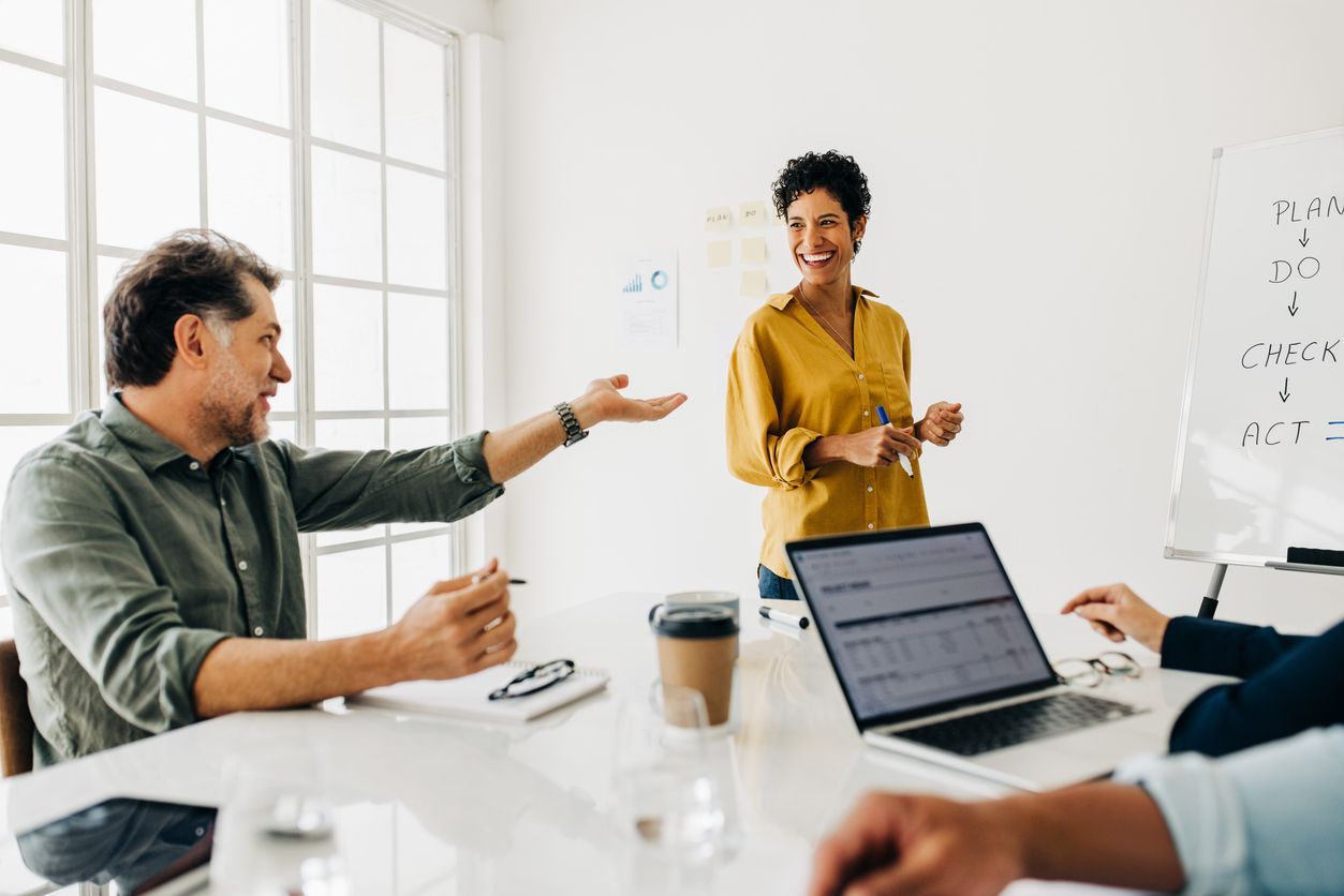 Business meeting: Woman in yellow shirt presenting; man gestures with hand, others at table. Bright room, whiteboards, laptop.