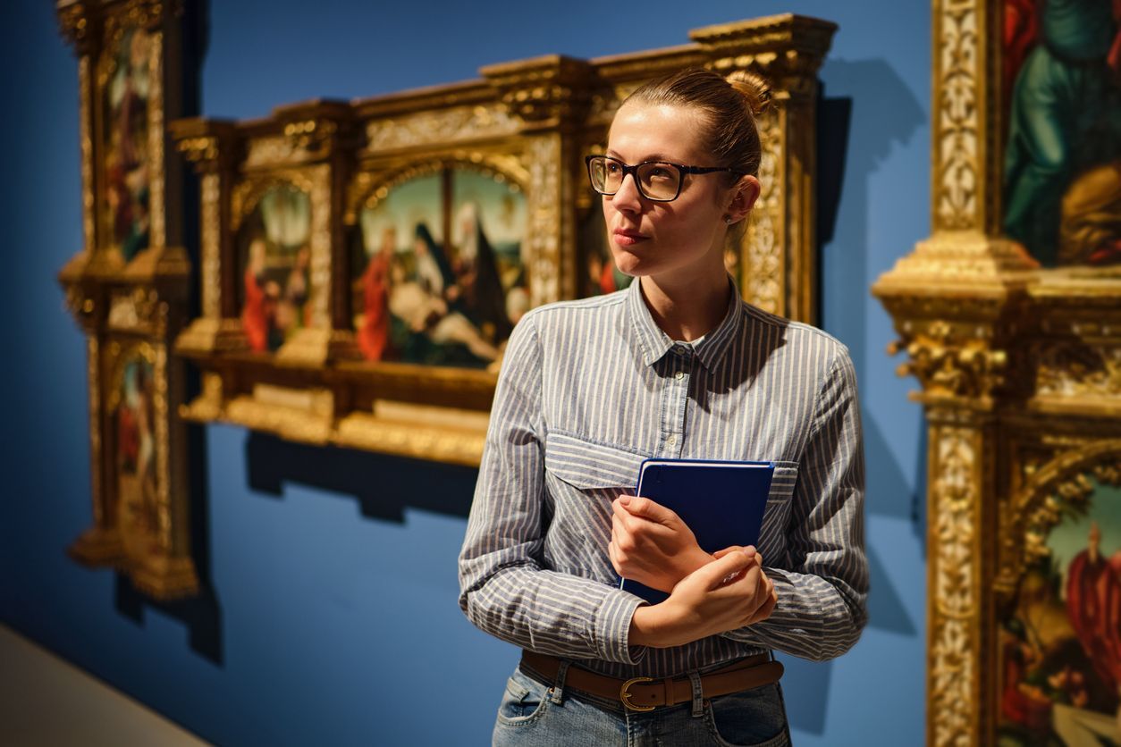 Person with glasses in a museum, looking at paintings, holding a blue book.