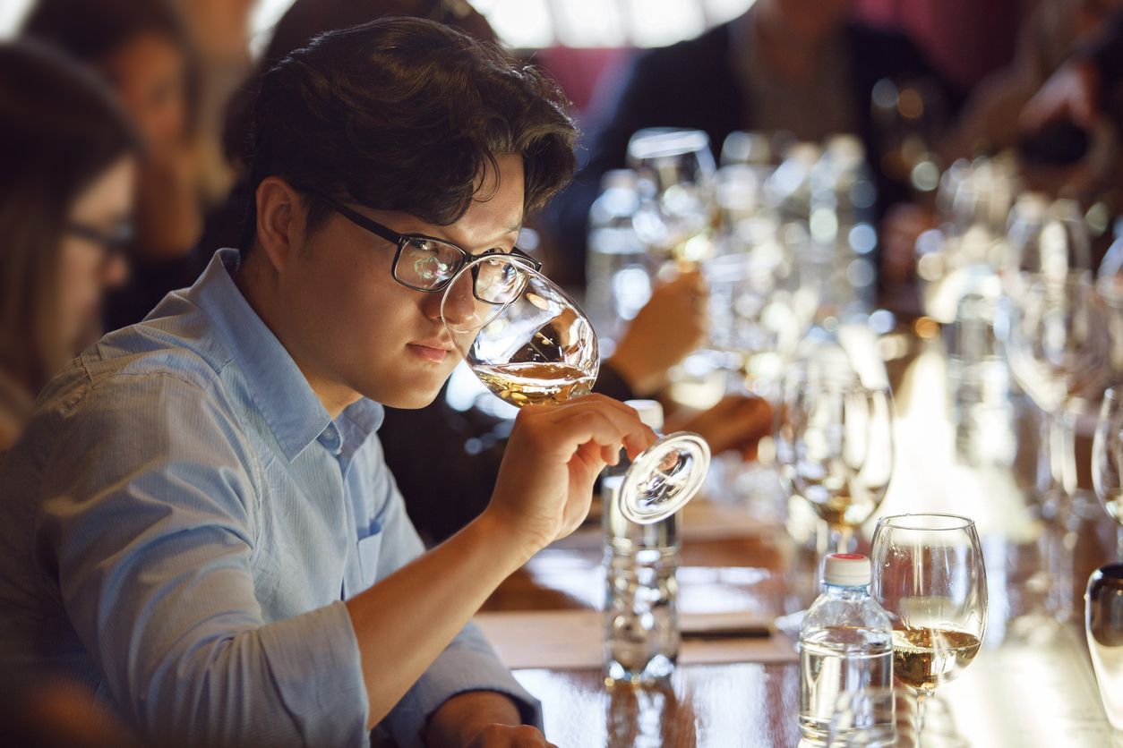 Man smelling wine in a tasting setting; glasses, bottles, and others in background.