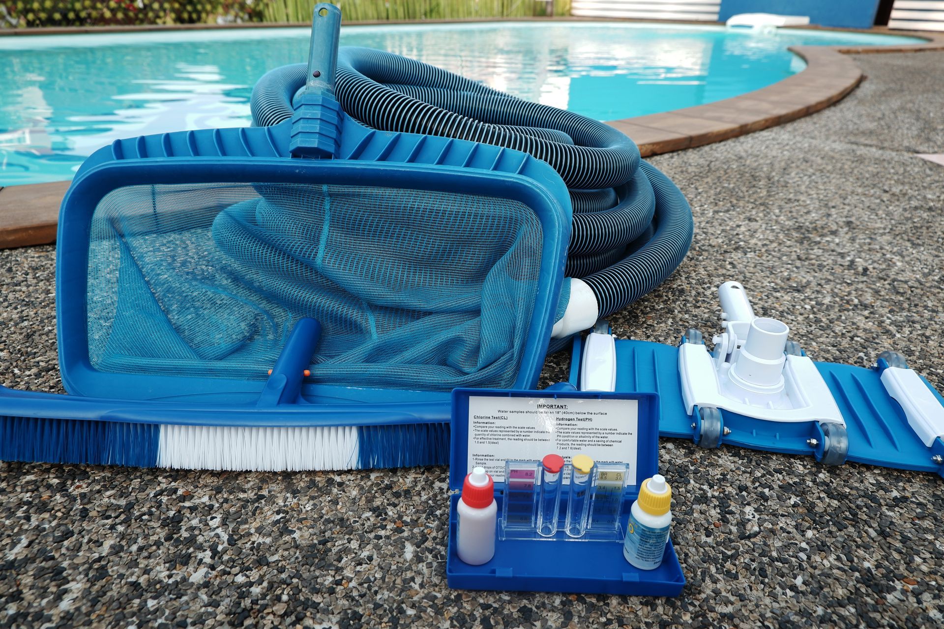 Pool cleaning tools and supplies laid out beside a swimming pool on a pebble deck