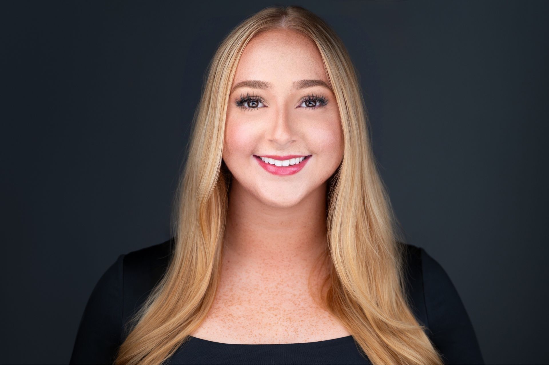 Woman with long blonde hair, smiling, in front of a dark gray background.