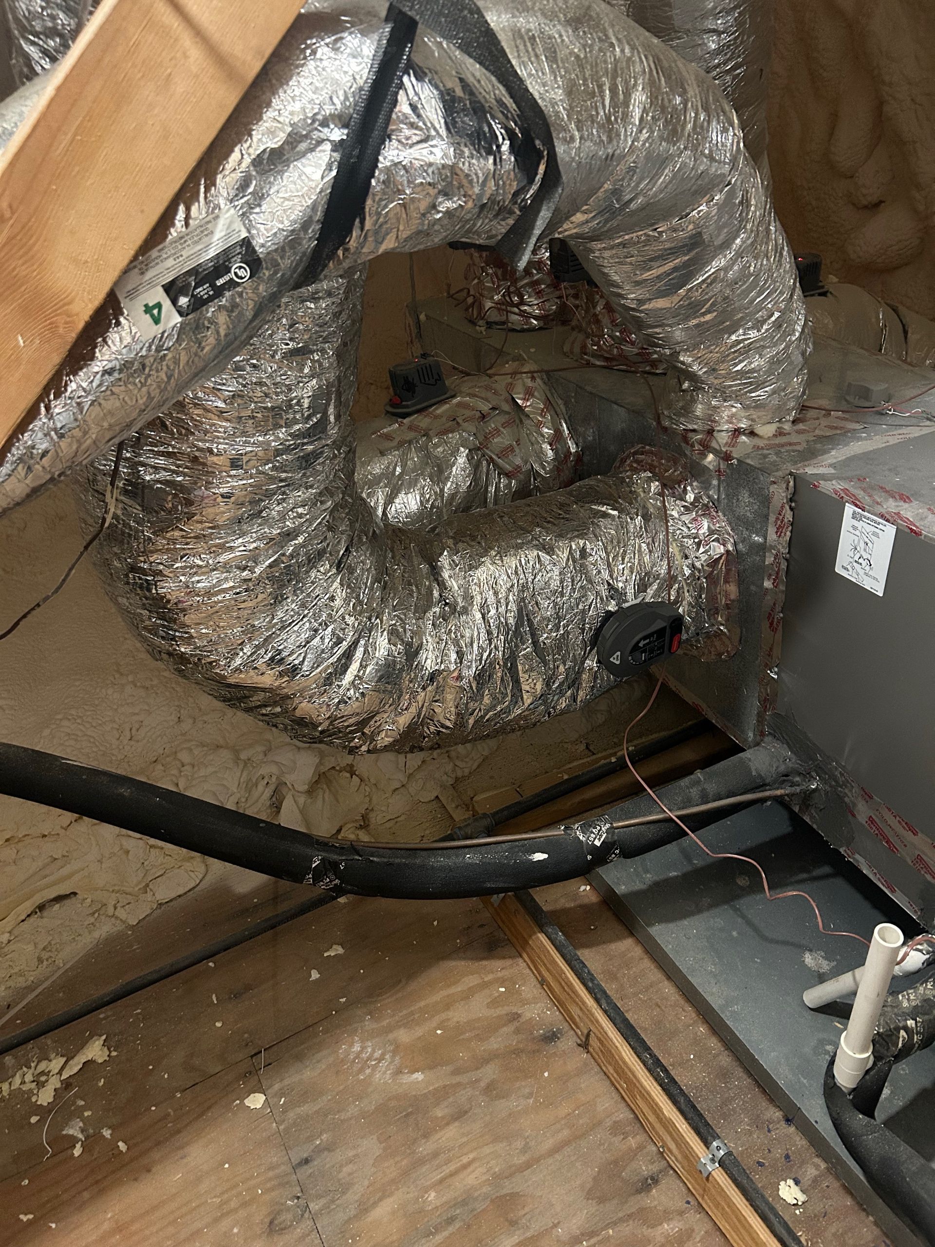 Silver insulated air ducting coiled in an attic space, near a black pipe and a unit.