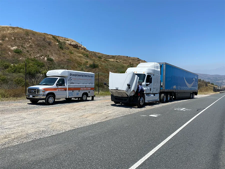 A white Amazon semi-truck with a raised hood and a repair truck parked on the side of a road.