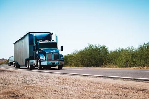 Blue semi-truck driving on a road with green bushes and blue sky.