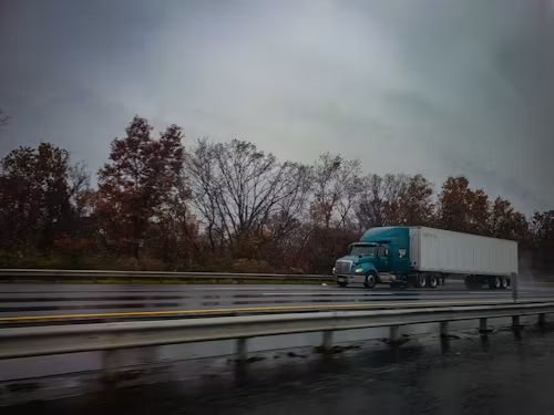 Blue semi-truck drives on a wet highway under a cloudy sky, with trees in the background.