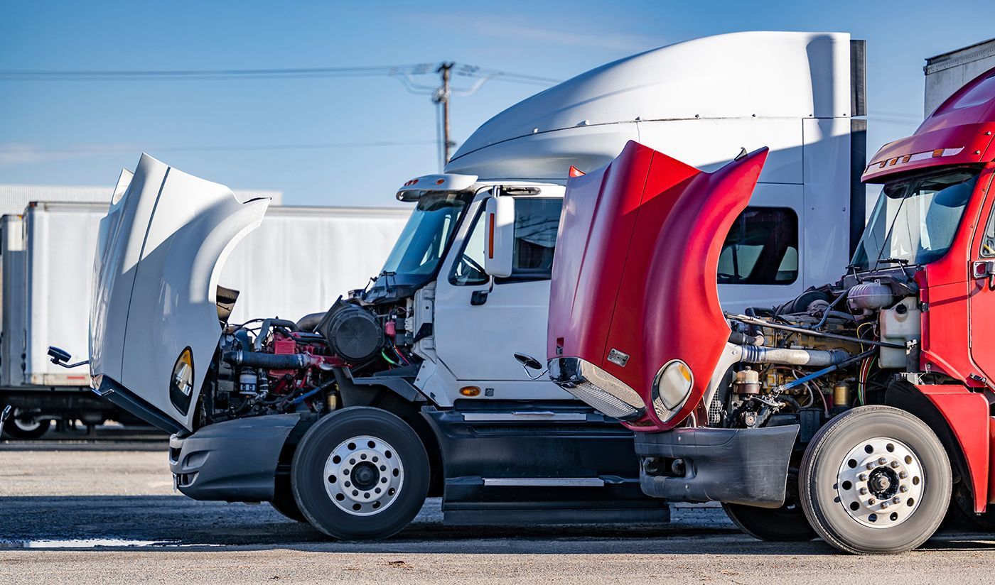 Semi-trucks with open hoods parked outside on a sunny day. White and red trucks are visible.