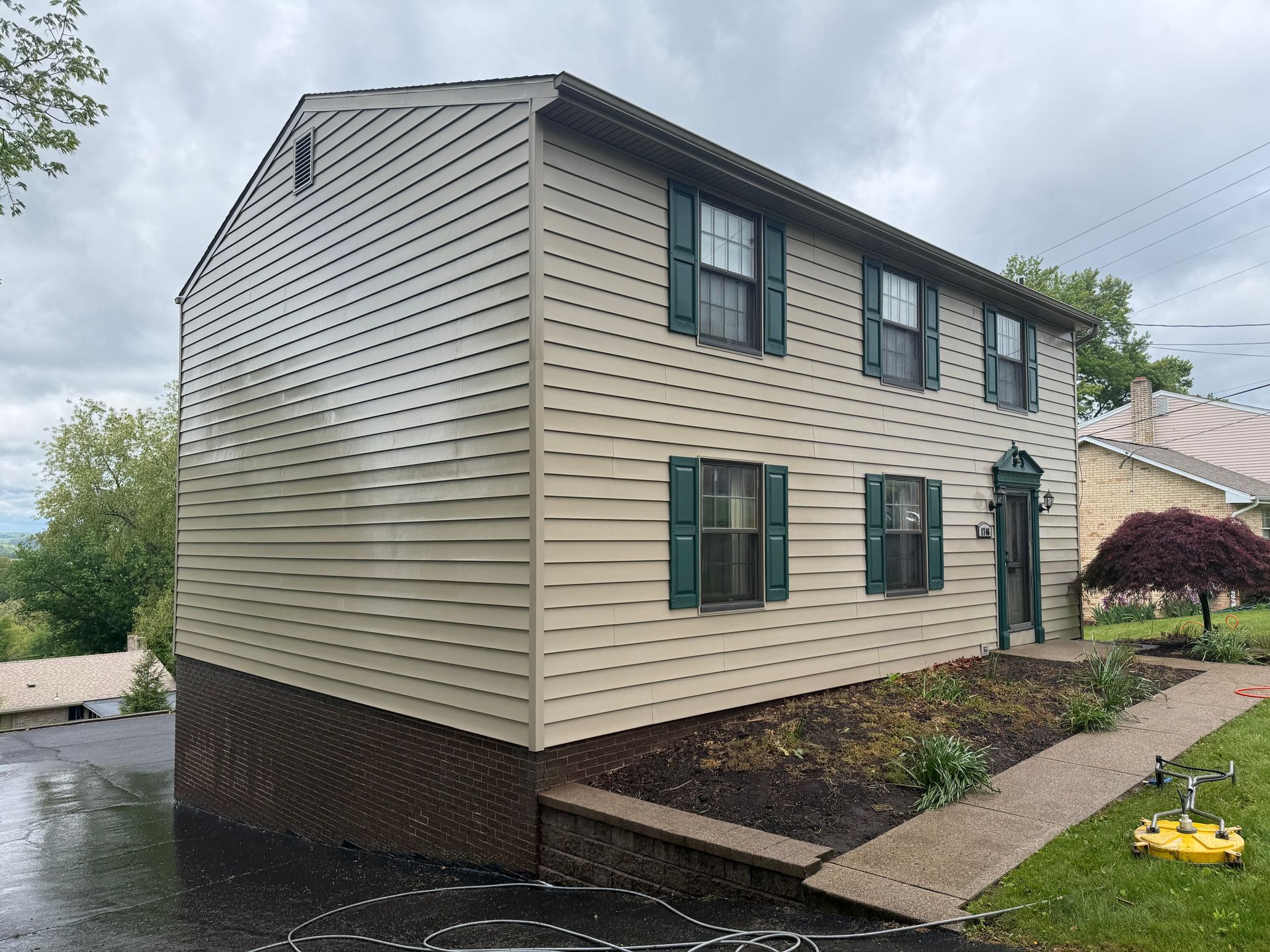 Two-story beige house with green shutters on a wet driveway after being pressure washed