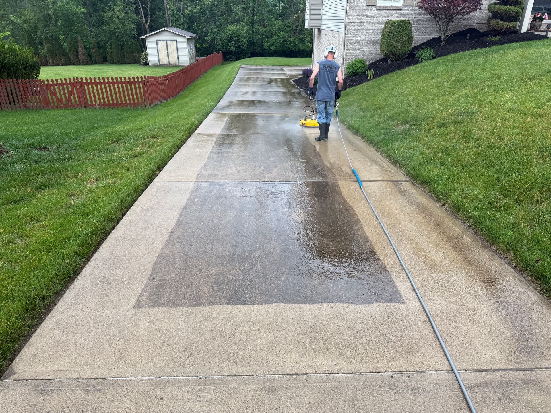Person power-washing a long concrete driveway beside a grassy lawn and stone house.
