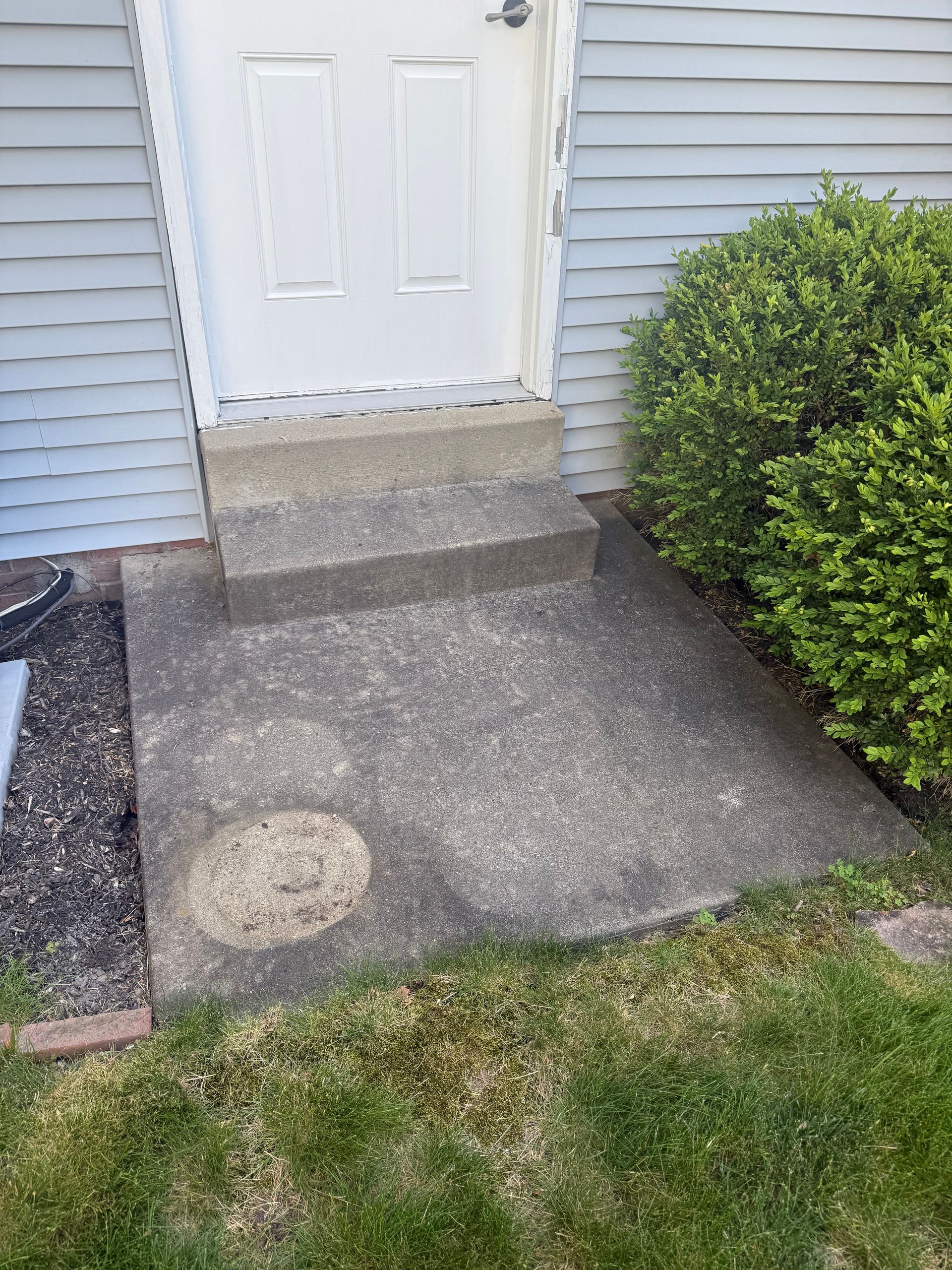 Front stoop with white door, concrete steps, and a small walkway beside a green bush.