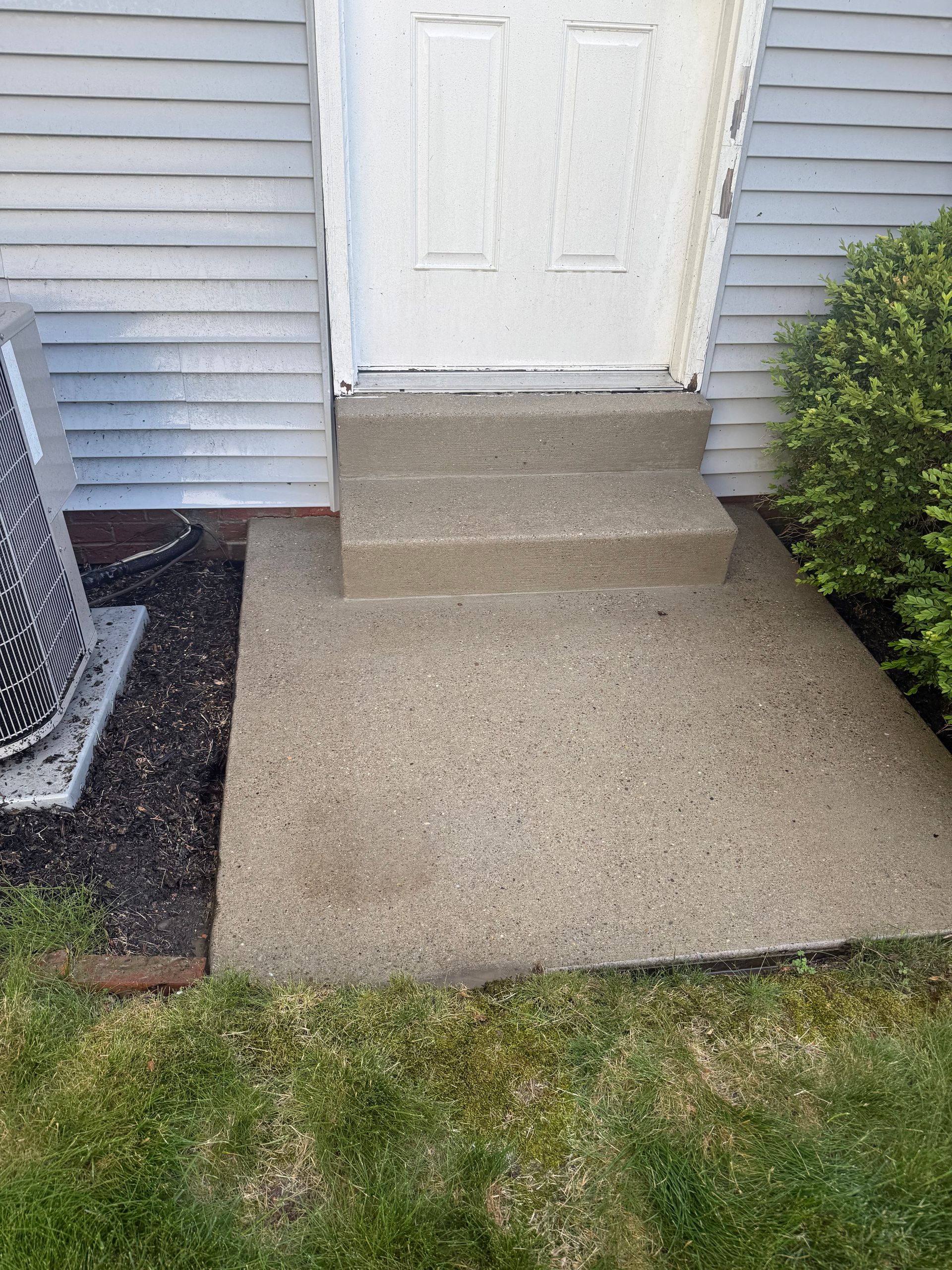 Front porch with concrete steps leading to a white door, bordered by siding, shrubs, and a small lawn.
