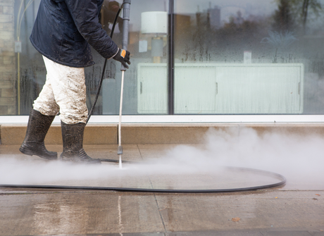 Worker pressure-washing a sidewalk beside a glass storefront, spraying water across the pavement