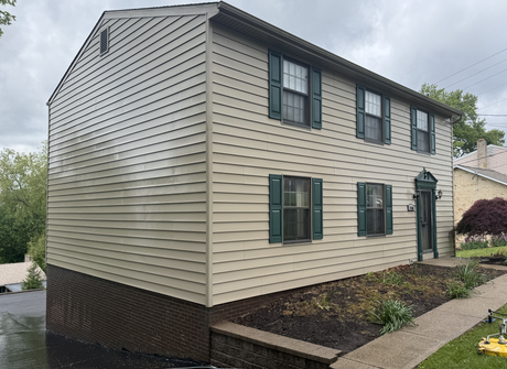 Two-story beige house with dark green shutters and a front walkway in a suburban neighborhood