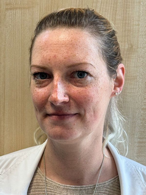 Frau mit heller Haut und hellem Haar, trägt einen weißen Kittel und blickt in die Kamera.