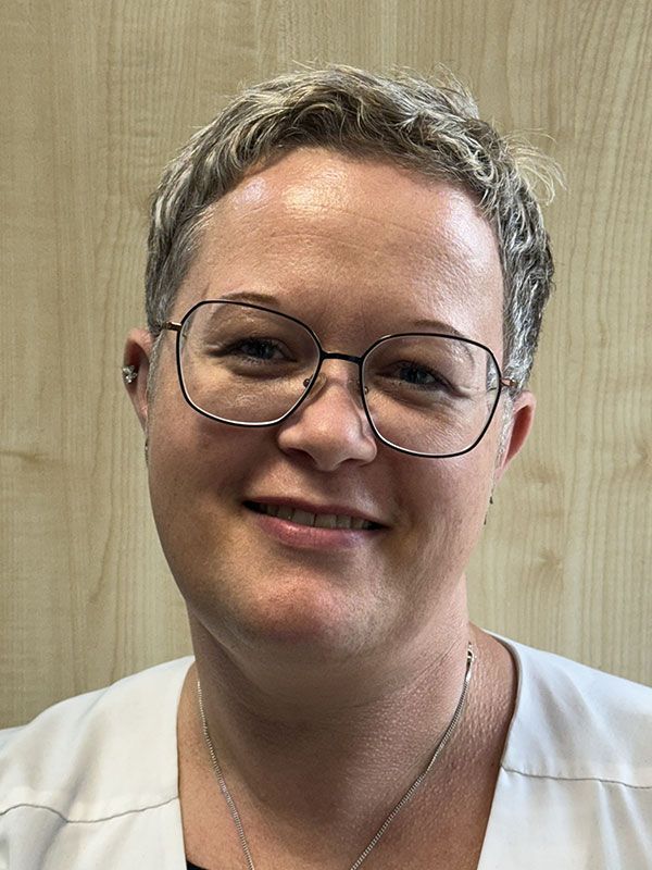 Frau mit kurzen Haaren, Brille und weißem Oberteil, lächelt vor einer holzgetäfelten Wand in die Kamera.
