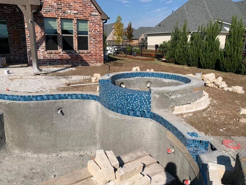 Backyard with unfinished concrete pool and raised spa, blue tile trim, near a brick house.