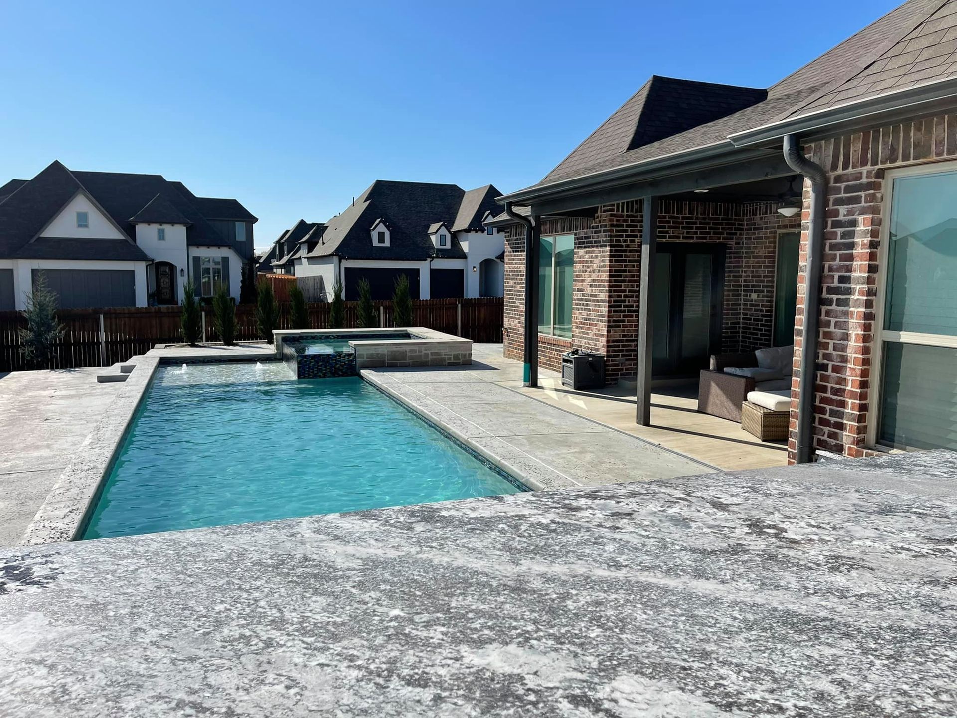 Backyard with a turquoise swimming pool beside a brick house and patio seating, surrounded by modern homes