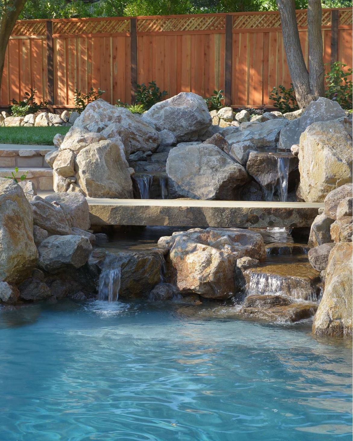 Rock waterfall flowing into a blue swimming pool, with wooden fence and boulders surrounding it.