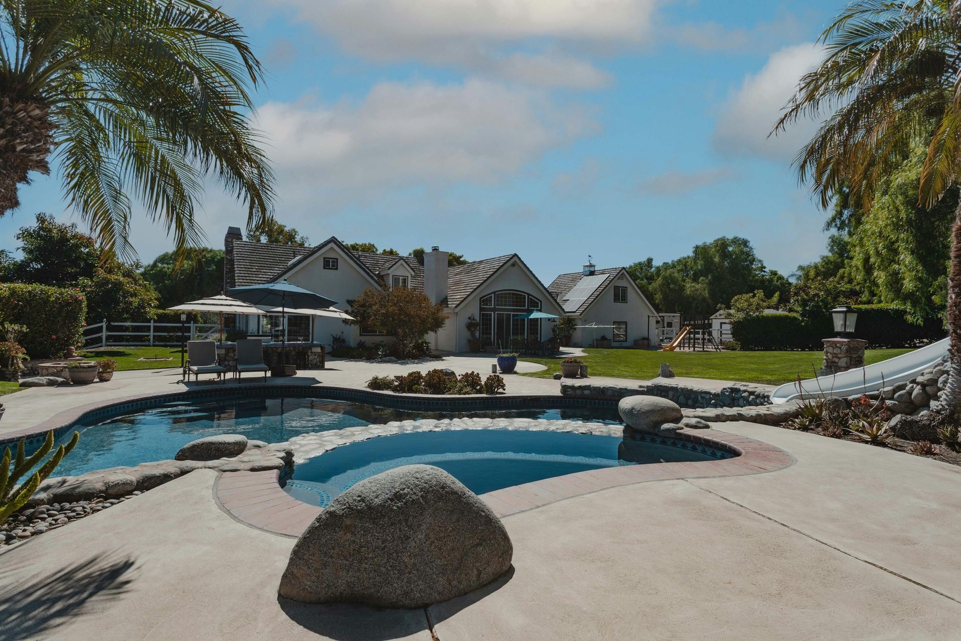 Backyard pool with palm trees, lounge chairs, and a landscaped patio beside houses under a blue sky