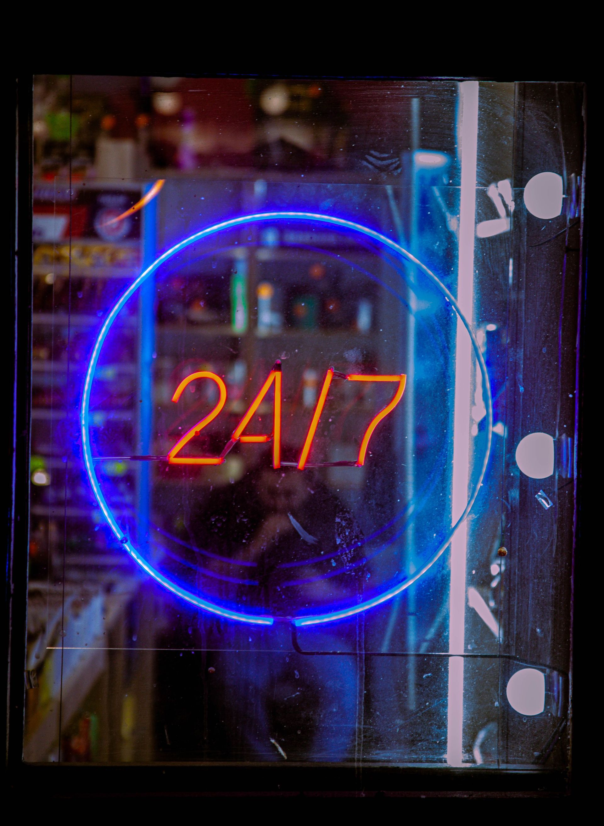 Neon “24/7” sign glowing orange inside a blue circle on a dark storefront window.