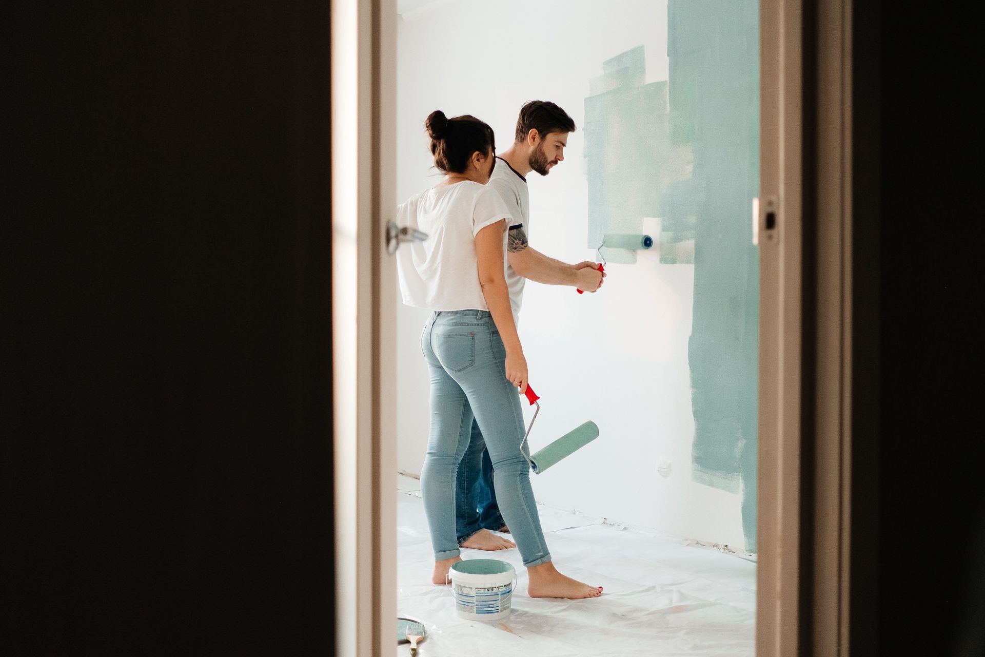 Two people painting a white wall, one using a roller in a bright room