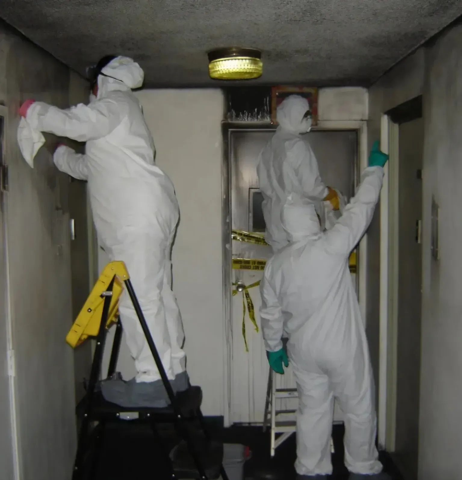 Two people in white coveralls paint a narrow room, one on a ladder and one on the floor.