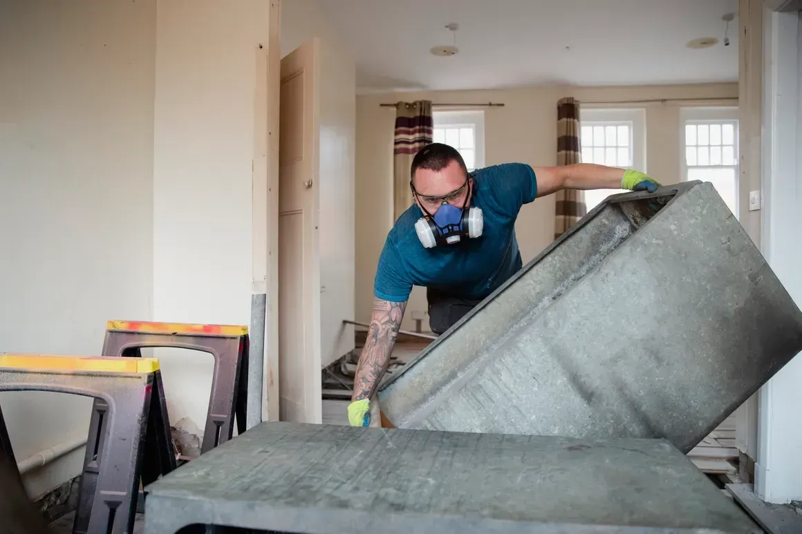 Worker in a respirator moving a large sofa in a bright, empty room during renovation