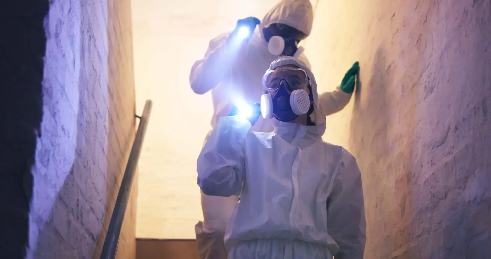 Two hazmat-suited workers with flashlights in a narrow stairwell, one at the top and one below.
