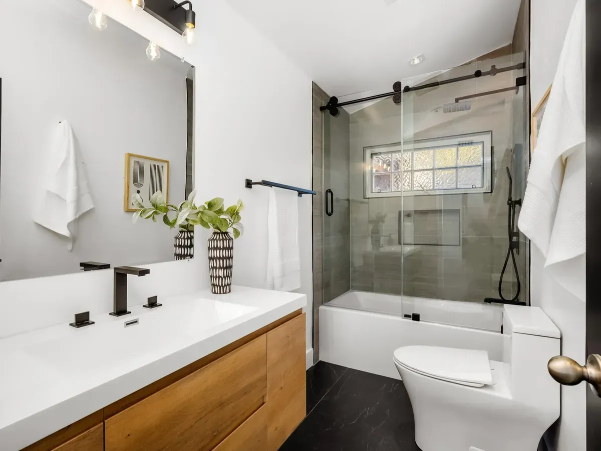 Modern bathroom with a wooden vanity, white countertop, large mirror, glass-enclosed tub, and a white toilet.