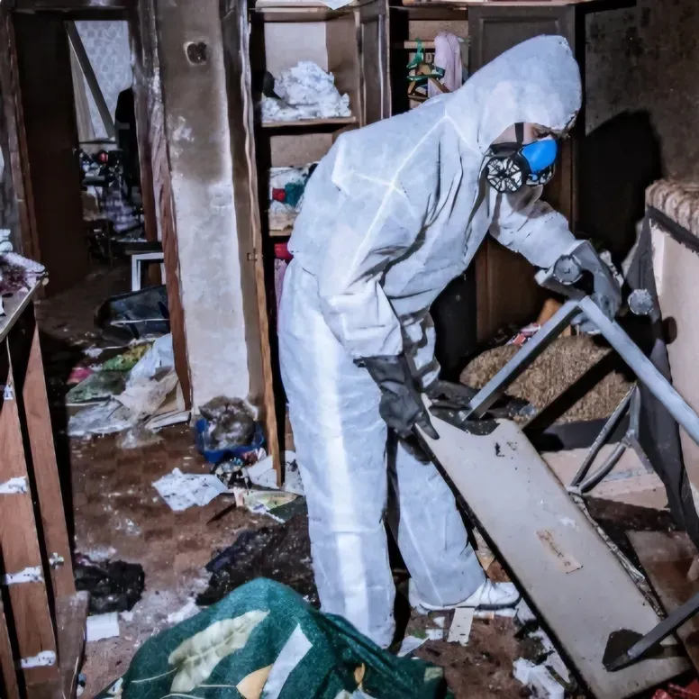 A person in a white hazmat suit and mask inspects a piece of furniture in a cluttered, debris-filled room.