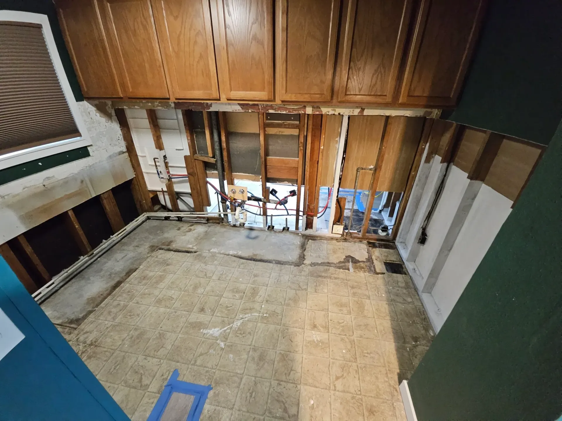 A kitchen undergoing renovation with removed countertops, exposed wall studs, visible plumbing, and upper wood cabinets.