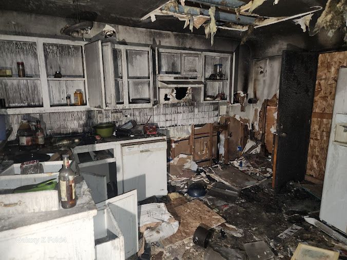 Burned, debris-strewn kitchen with charred cabinets, broken appliances, and soot-covered walls