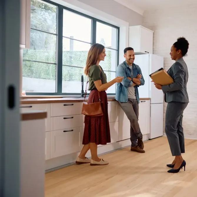 Three people talking in a bright kitchen, one holding a clipboard and two smiling near a window.
