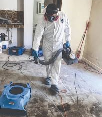Worker in white coveralls using a floor cleaning machine on a dusty room floor.
