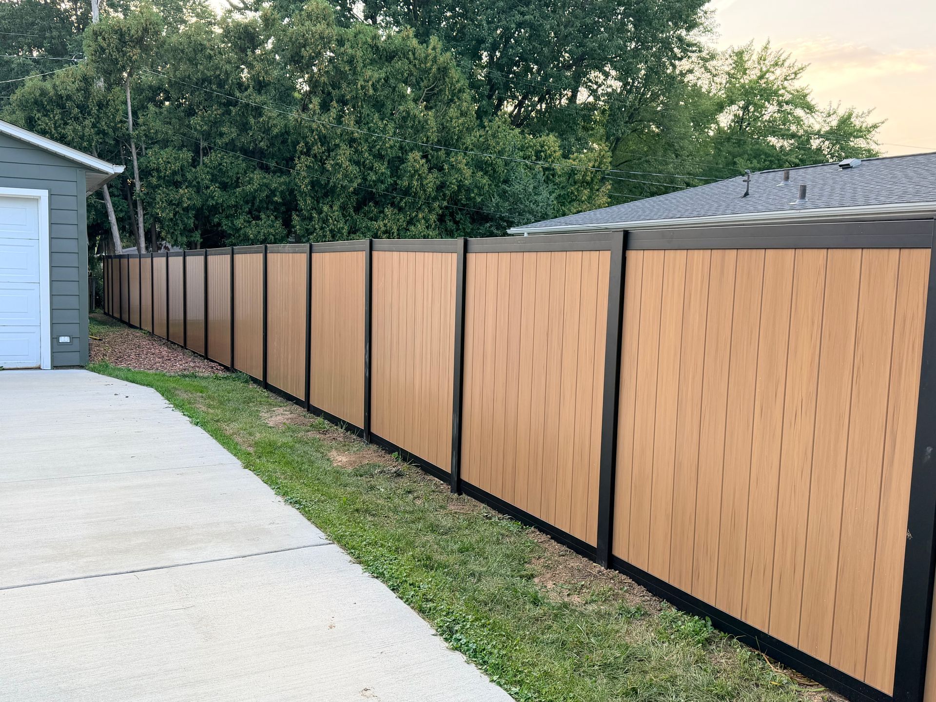 Tan-and-black privacy fence lining a grass strip beside a driveway and house.