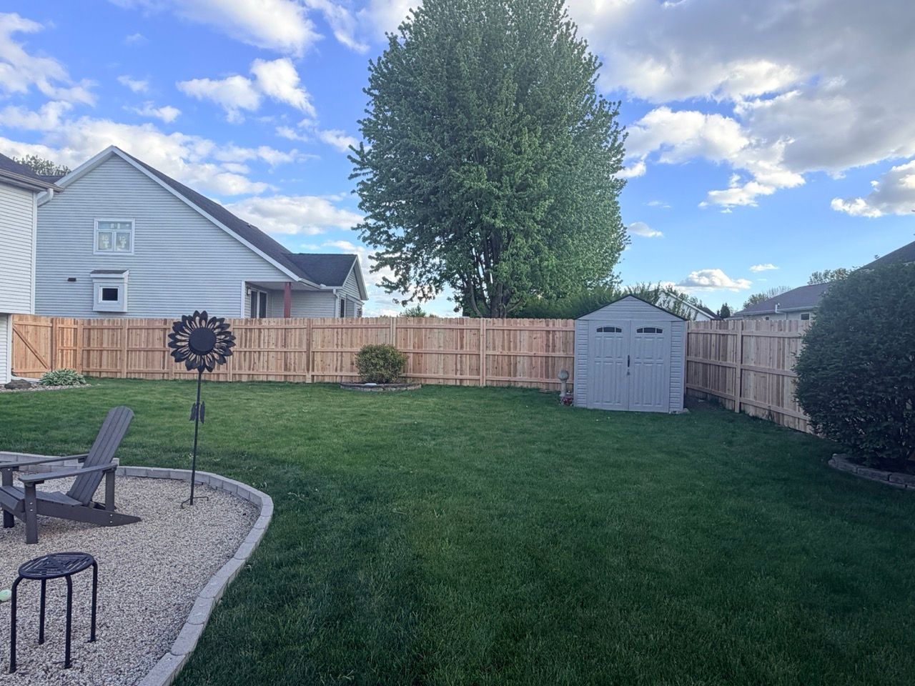 Backyard with green lawn, wooden fence, shed, patio chairs, and a large tree under a partly cloudy sky