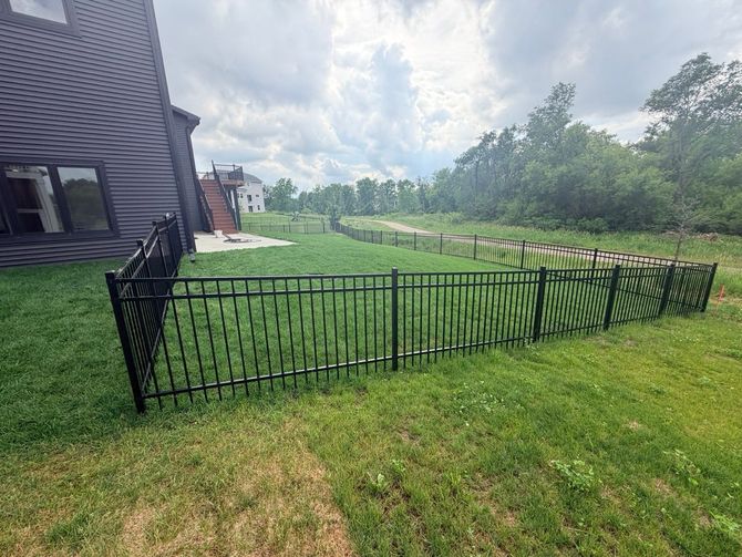 Black metal fence enclosing a green lawn beside a house, with trees and sky in the background