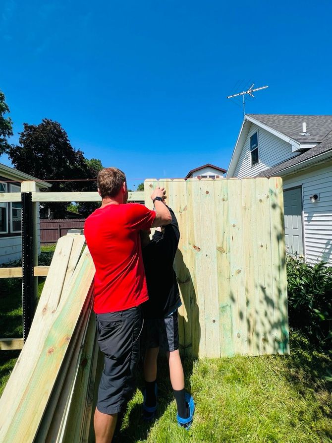 Two people painting a wooden fence in a sunny backyard, with a ladder leaning nearby.