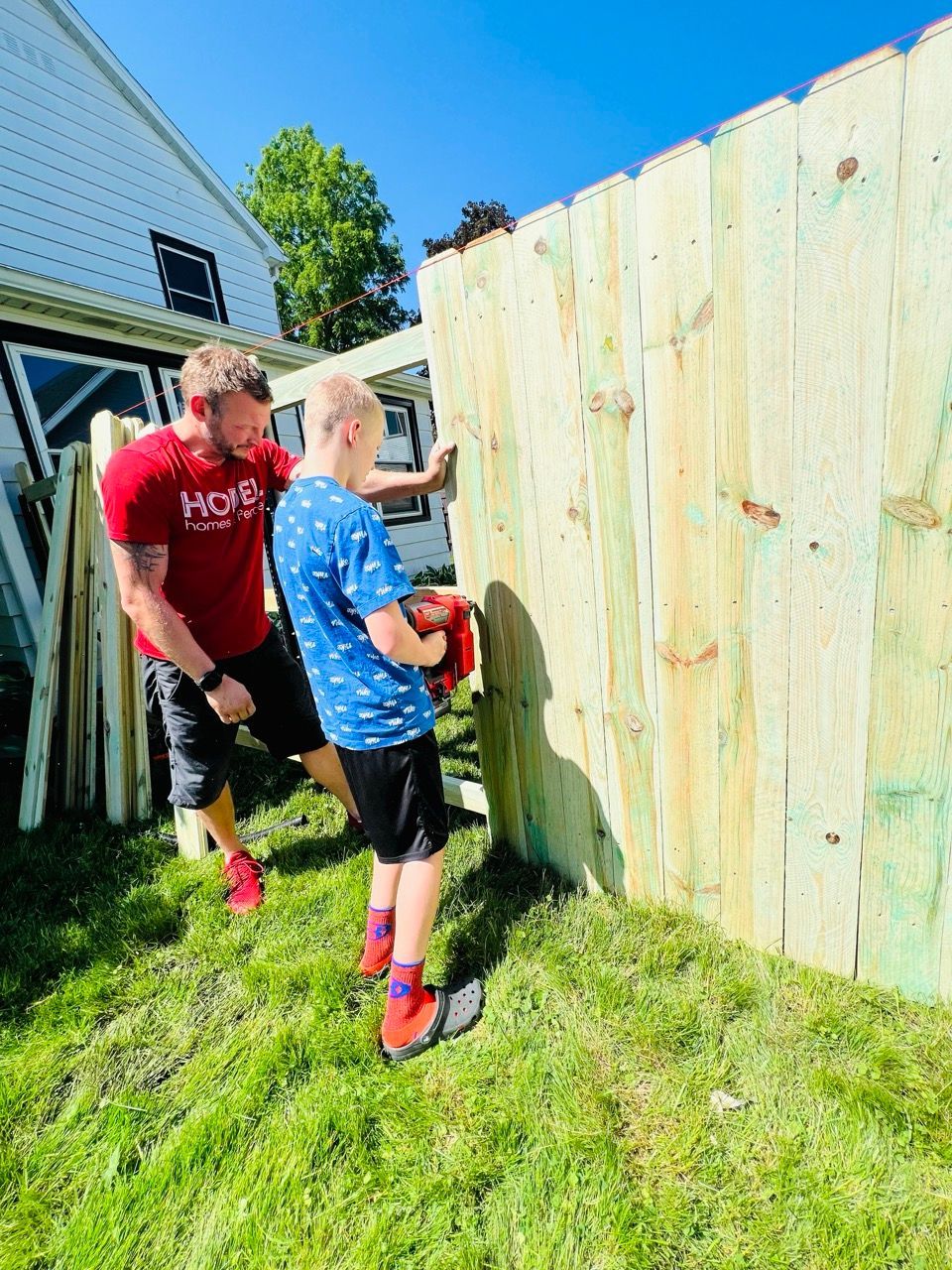 Two people working beside a tall wooden fence on a sunny grassy yard