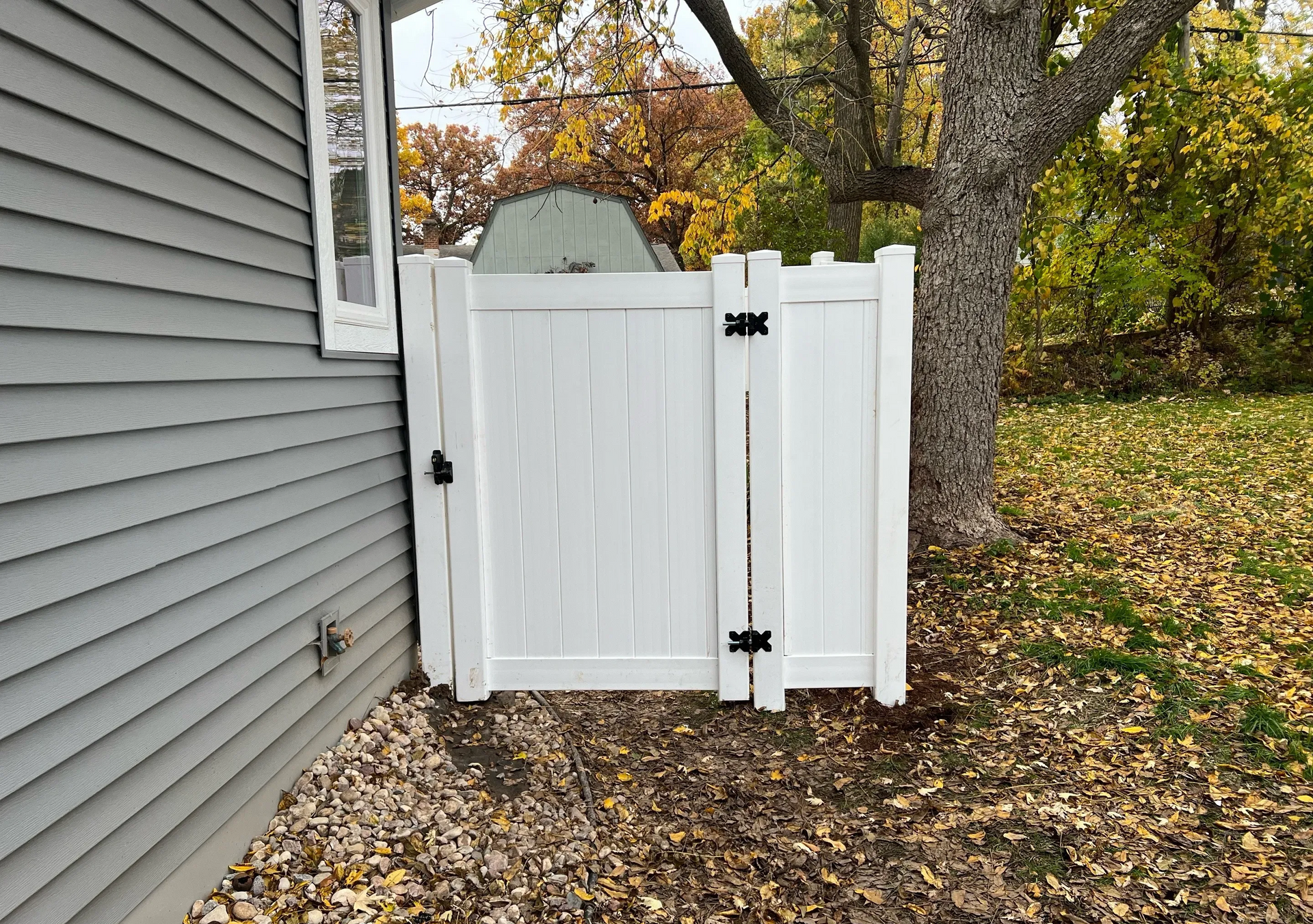 White vinyl gate beside gray house, with autumn leaves and trees in the yard