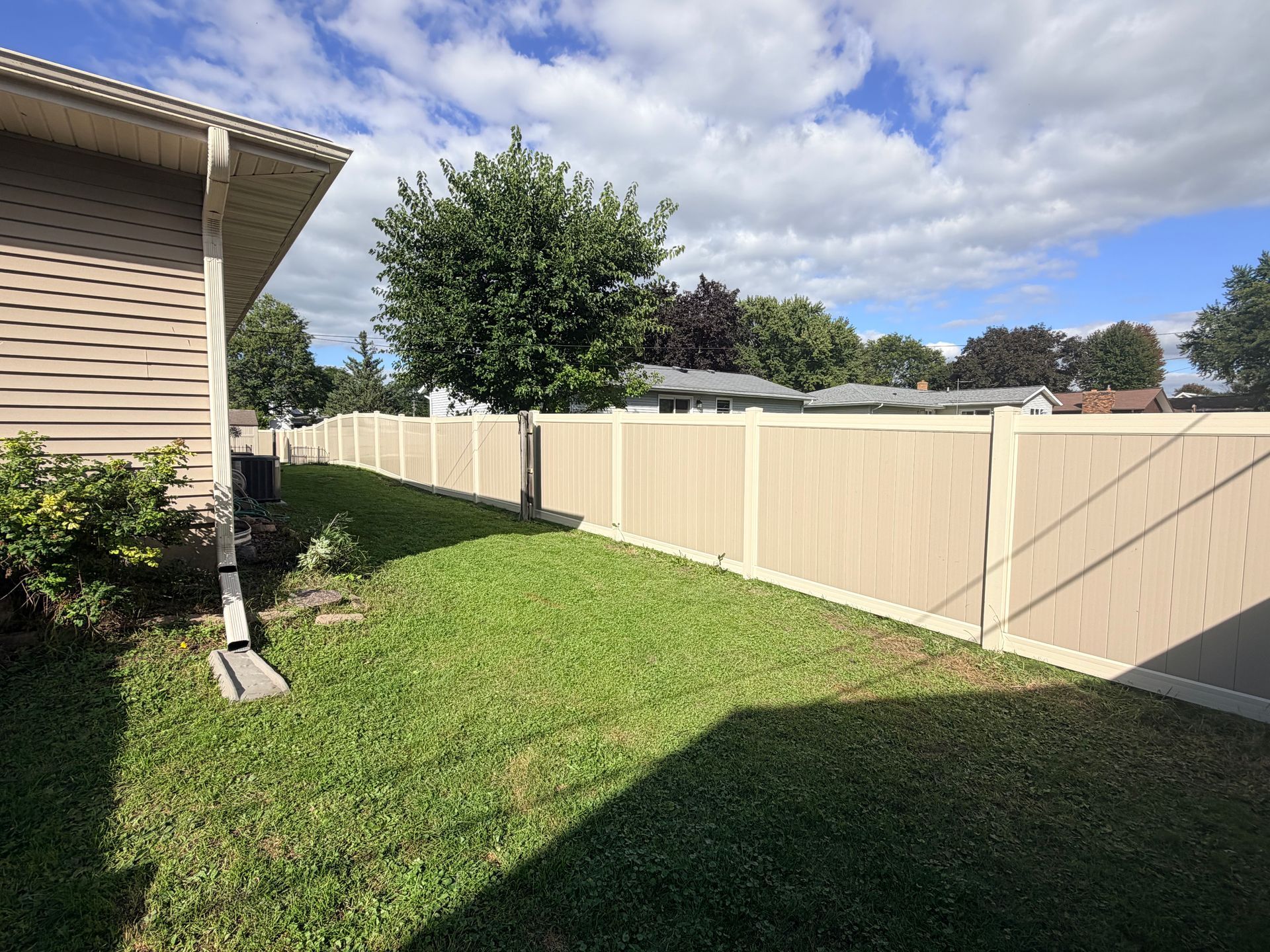 Backyard with green lawn, beige fence, and partly cloudy sky beside a house