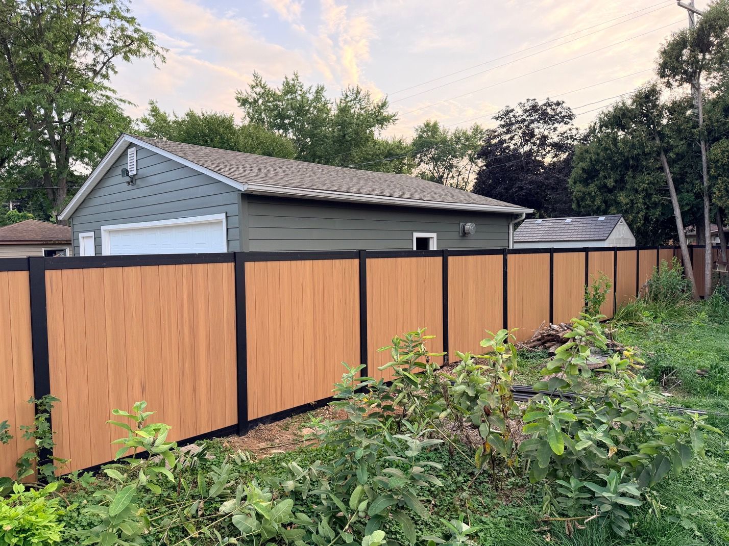 Wooden privacy fence in a yard beside a gray garage, with dense greenery in the foreground.