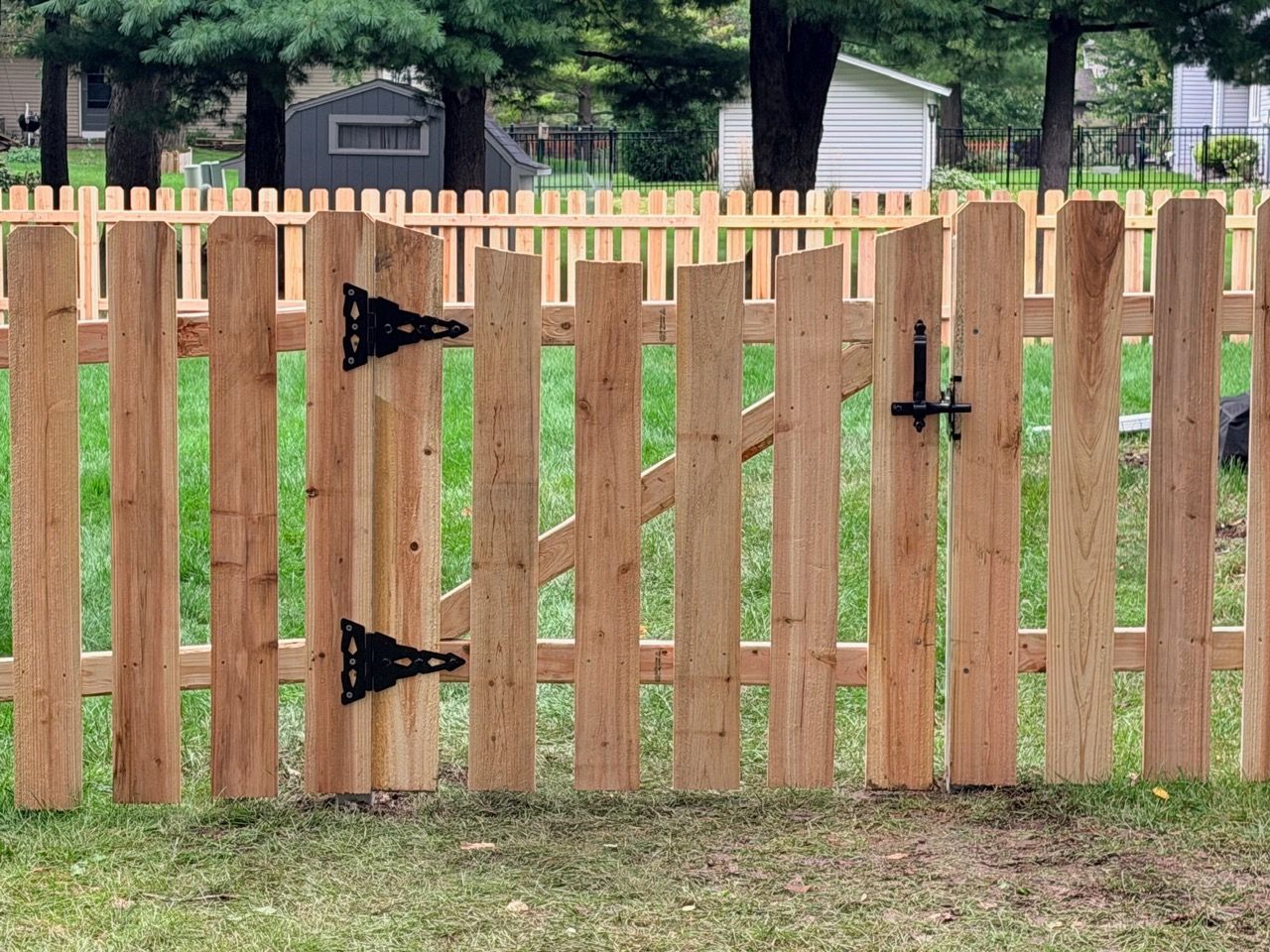 Wooden backyard gate with black hinges and latch in a fenced yard.