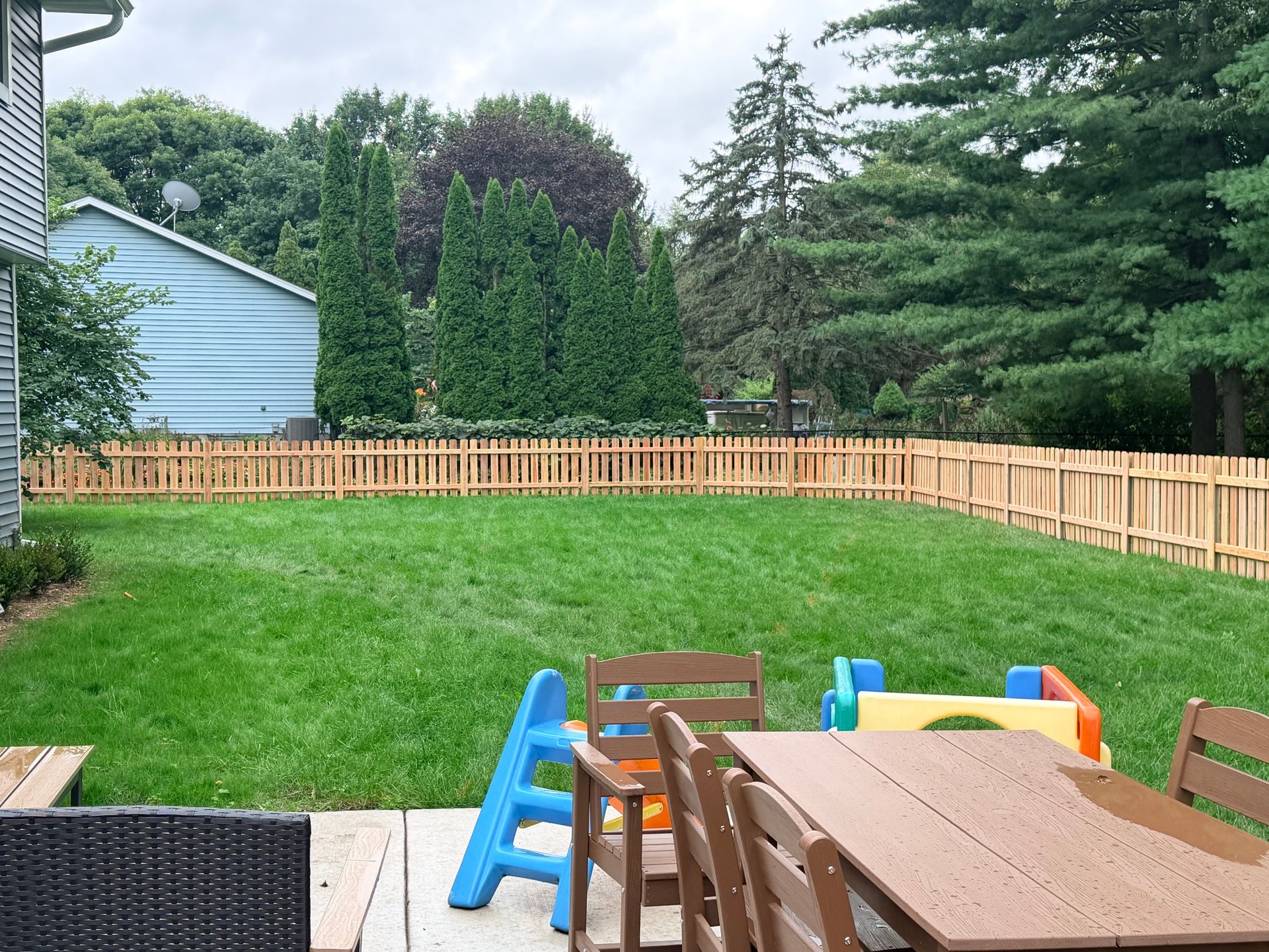 Backyard with green lawn, wooden fence, trees, and colorful children’s toys on a patio table