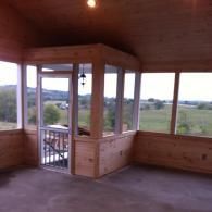 Interior view of a rustic, wooden-walled screened-in porch featuring a screen door and large windows overlooking a field.