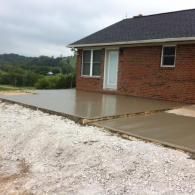 A newly poured concrete patio attached to a red brick house exterior, with gravel in the foreground.