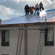 A group of construction workers in hard hats install blue roofing underlayment on a building with scaffolding and ladders.