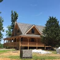 A two-story wooden cabin with a wraparound porch, a gray shingled roof, and surrounding trees under a clear blue sky.