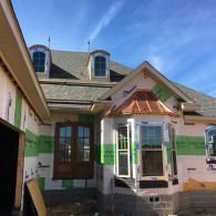 A house under construction with a copper-roofed bay window, dark wood doors, and visible green sheathing under a blue sky.